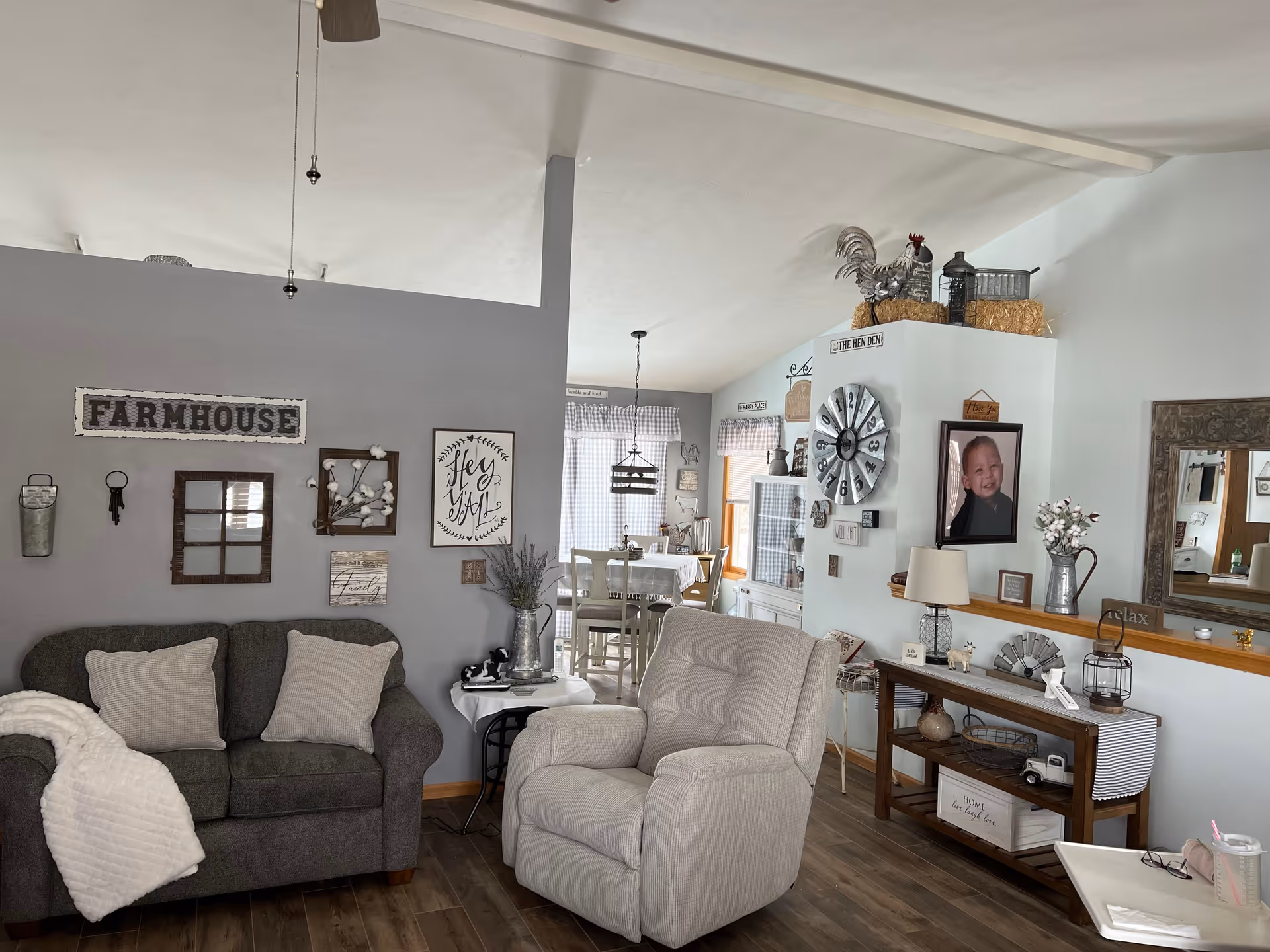A cozy living room area with a dark gray loveseat adorned with two light-colored pillows and a white throw blanket. Next to it is a light beige recliner chair. The walls are decorated with farmhouse-style signs and framed art. In the background, there is a dining area with a table and chairs, and various rustic decorations including a large clock and a rooster figurine on a shelf. The floor is wooden, and the overall decor has a warm, farmhouse aesthetic.