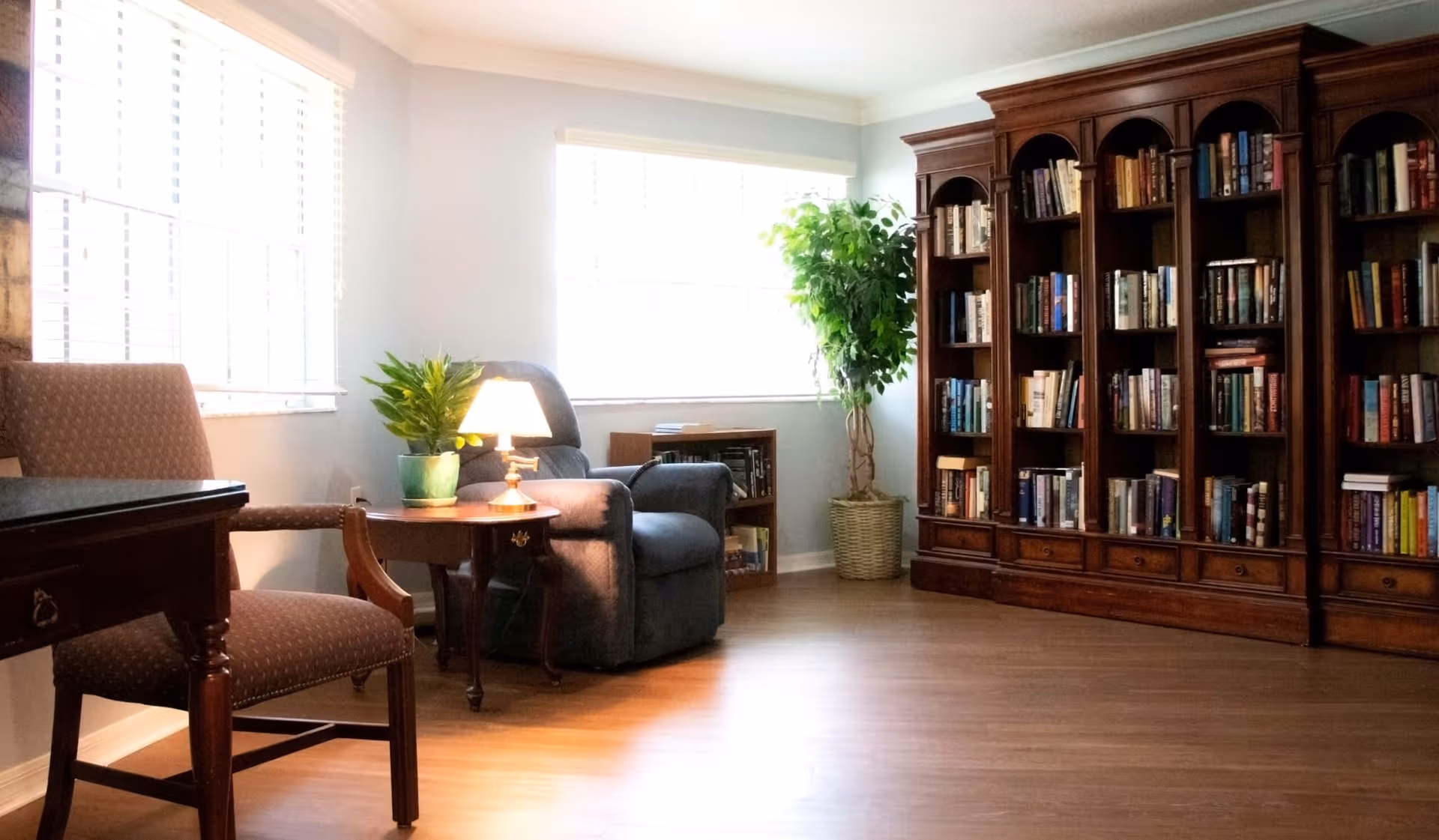 A cozy reading area in a senior living facility featuring a large wooden bookshelf filled with books, a comfortable recliner chair, a wooden side table with a small lamp and a potted plant, another chair, and large windows letting in natural light.