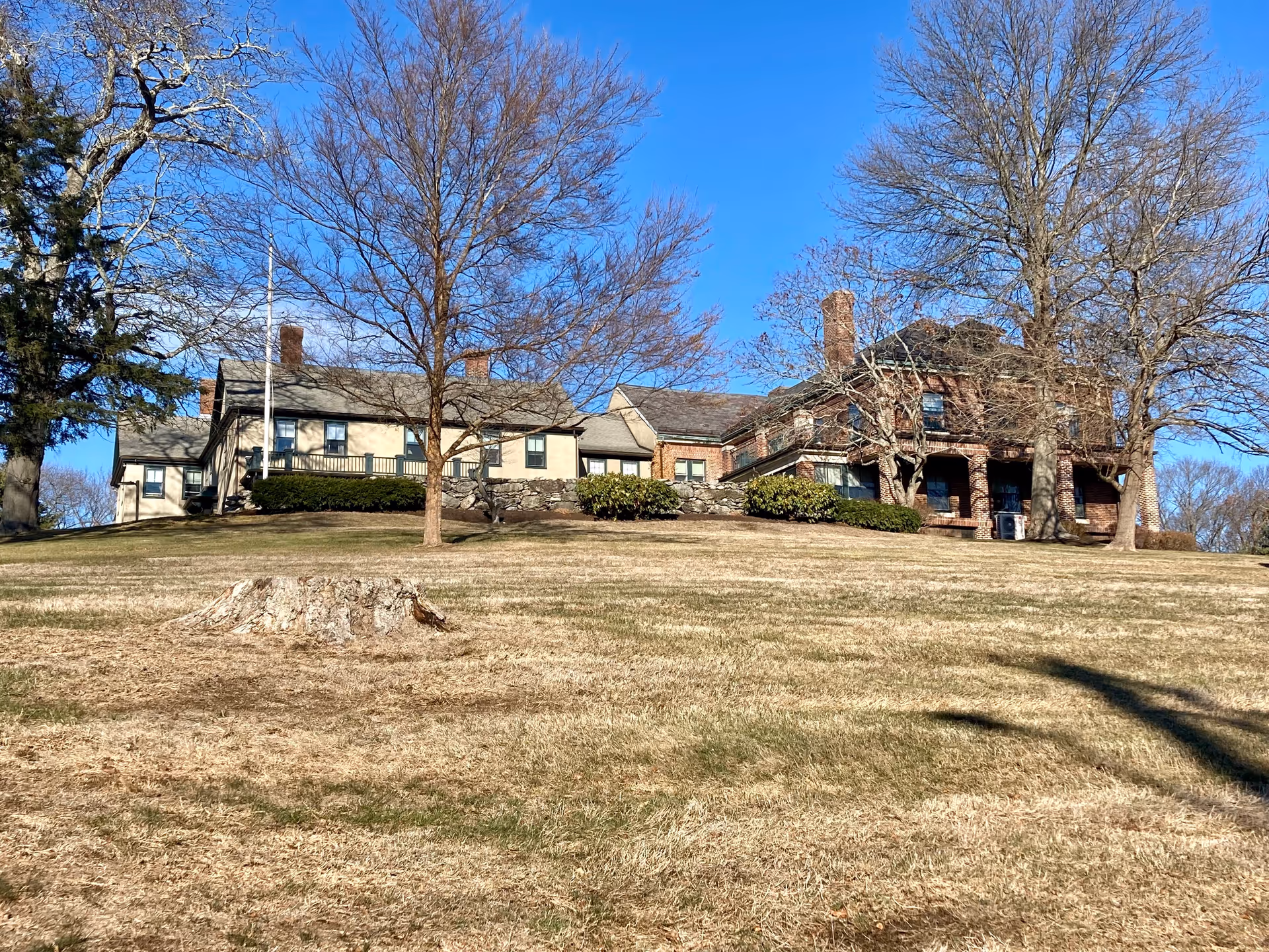 A large senior living facility building situated on a grassy hill with several leafless trees around it under a clear blue sky. The building has a combination of brick and light-colored siding with multiple chimneys and windows.