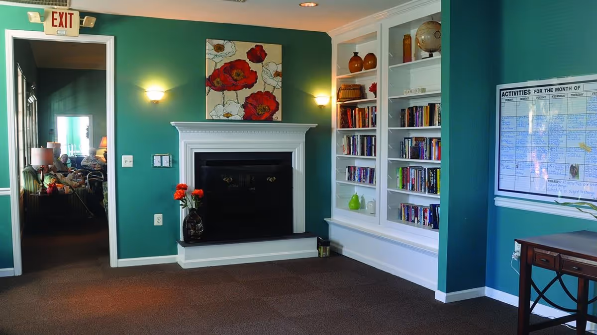 A cozy senior living common area with teal walls, a white fireplace with a vase of red flowers on the hearth, built-in white bookshelves filled with books and decorative items, a framed activity schedule on the wall, and an open doorway leading to another room where seniors are seated.
