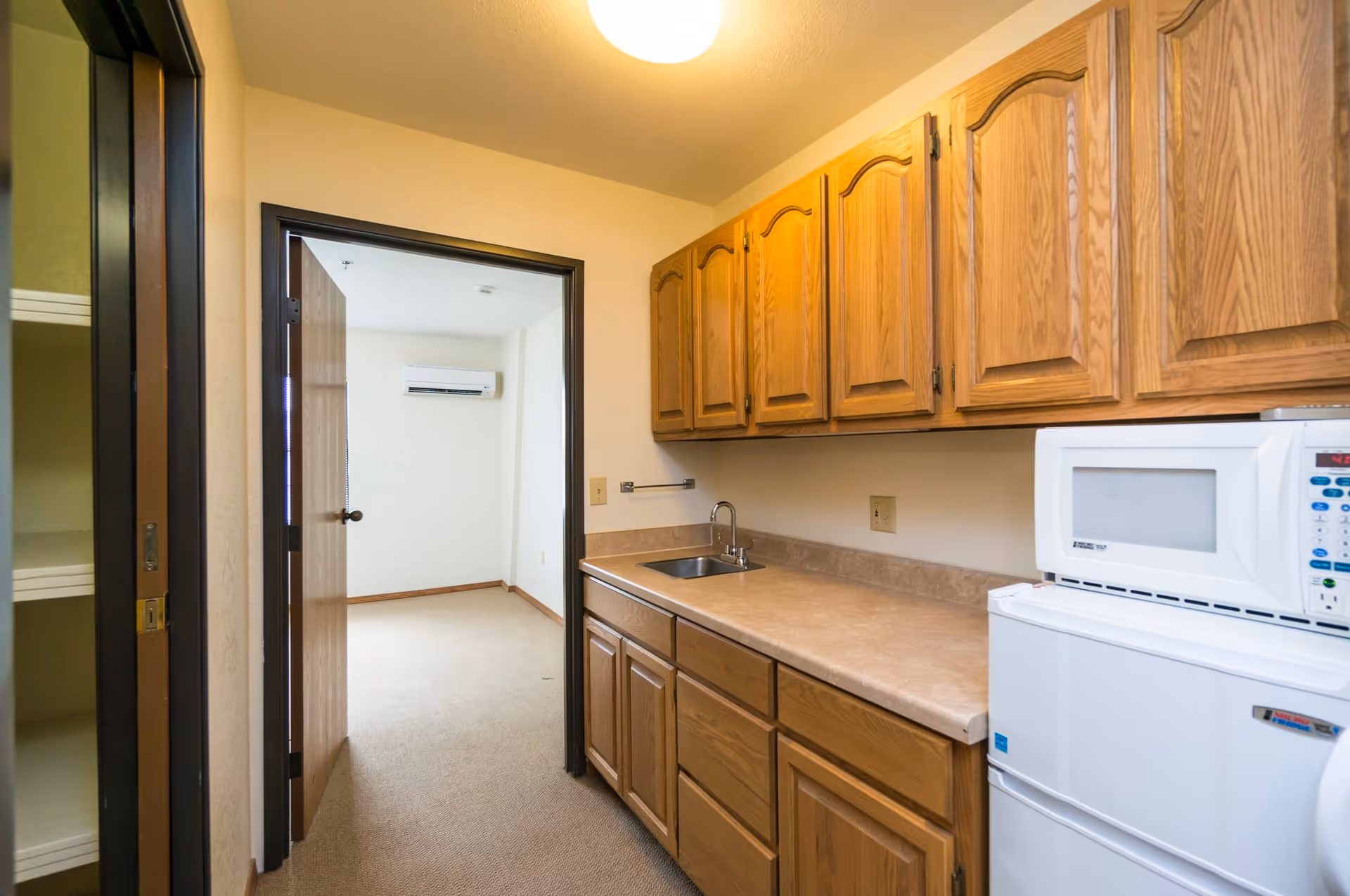 Small kitchenette area with wooden cabinets, a countertop with a sink, a microwave on top of a mini refrigerator, and an open door leading to an adjacent room with a wall-mounted air conditioning unit.