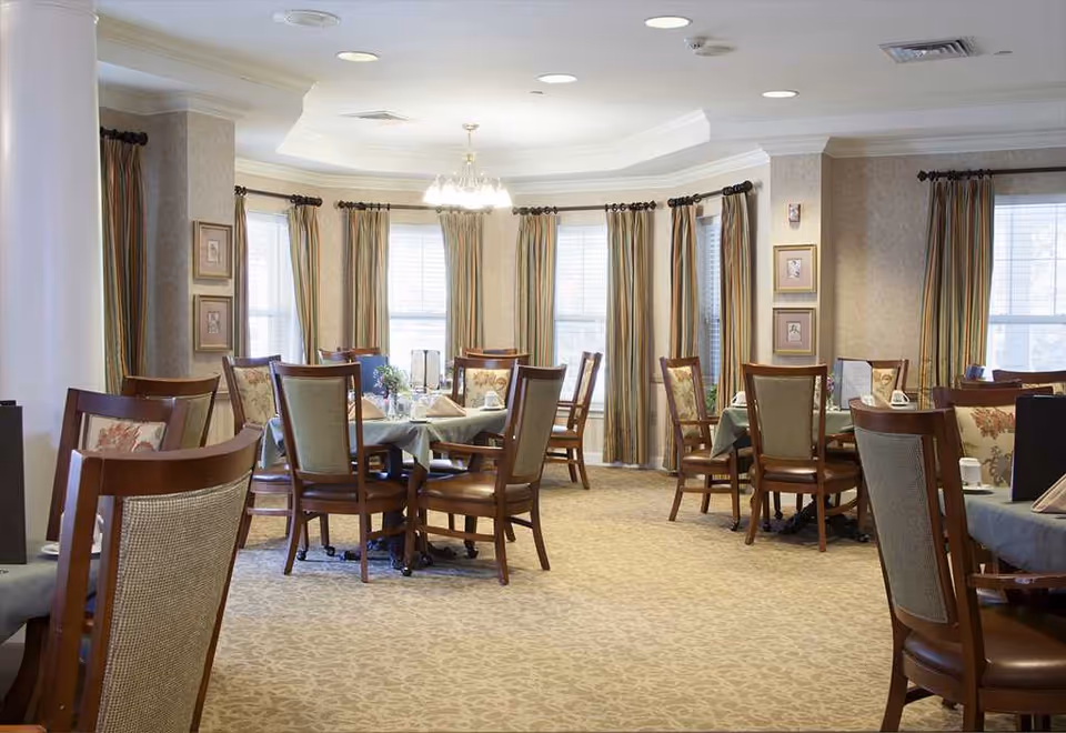 A bright dining room with round tables set for meals, wooden chairs, tall windows with curtains, and a central chandelier.