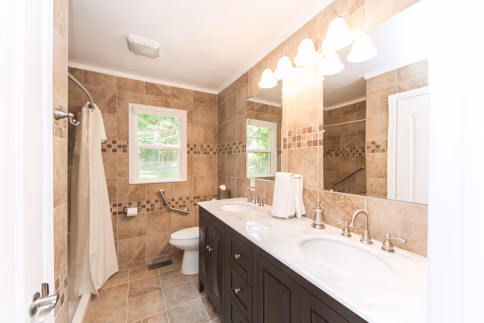 Well-lit bathroom with a double-sink vanity and large mirror, tiled walls and floor, a shower with curtain, and a window.
