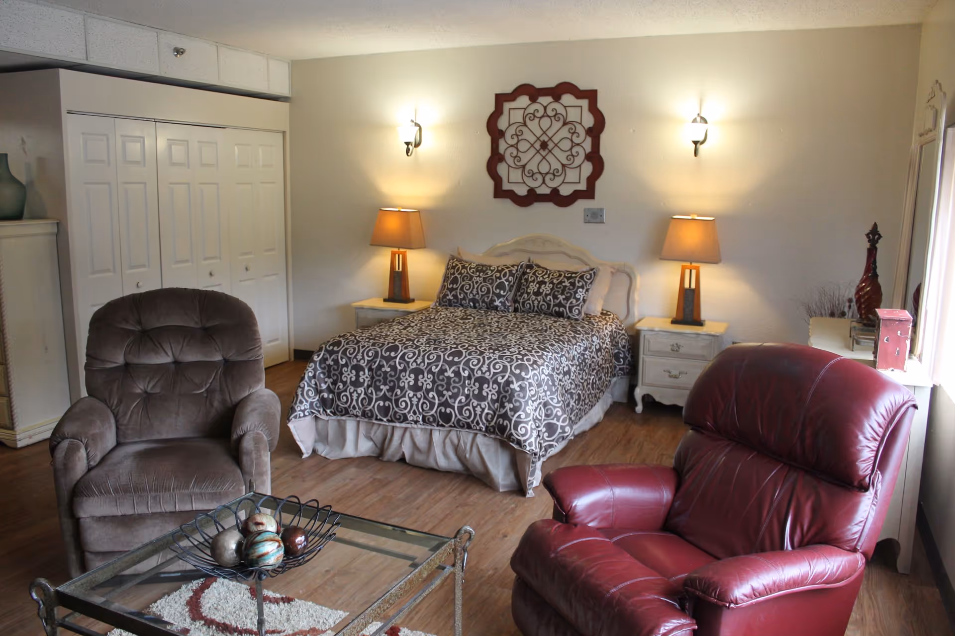 A cozy bedroom with a bed covered in a patterned bedspread, two bedside tables each with a lamp, a decorative wall piece above the bed, a brown upholstered armchair, a red leather recliner, and a glass coffee table with decorative balls on it. The room has wooden flooring and a closet with white sliding doors.