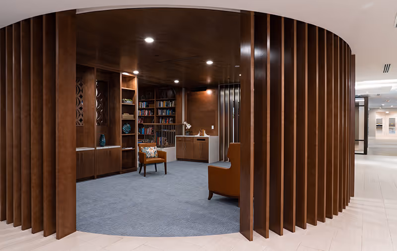 A modern, circular lounge area with wooden vertical slats forming a partial enclosure. Inside, there are built-in wooden bookshelves filled with books and decorative items, a small cabinet with a sink, and two upholstered chairs on a blue carpeted floor. The surrounding area has light-colored tile flooring and recessed ceiling lights.