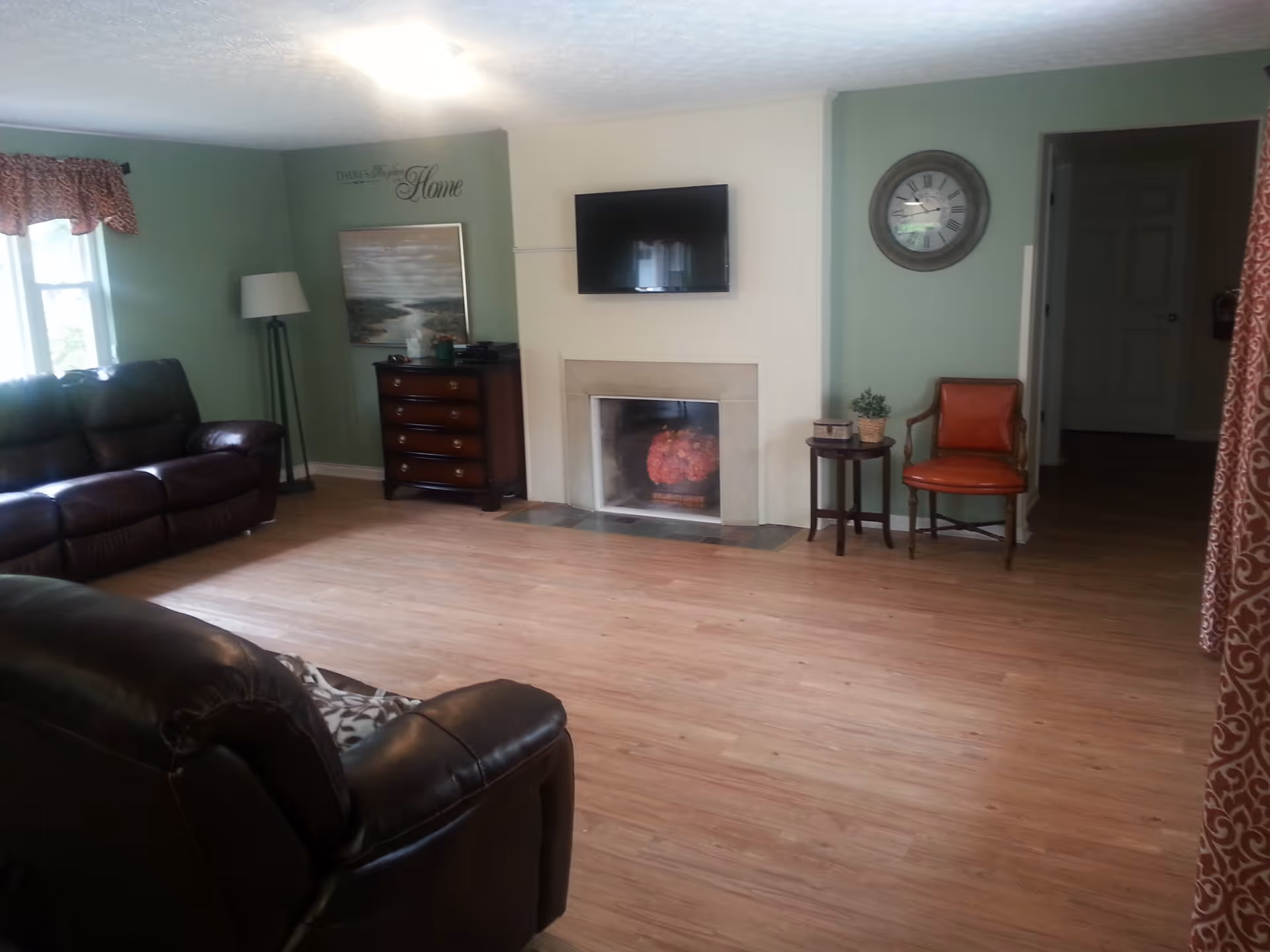 A cozy living room with light green walls and wood flooring. The room features two dark brown leather sofas, a fireplace with a TV mounted above it, a wooden chest of drawers with a lamp and decorative items, a round side table with a small plant, and an orange cushioned chair. There is a window with a patterned valance on the left and a wall clock on the right.