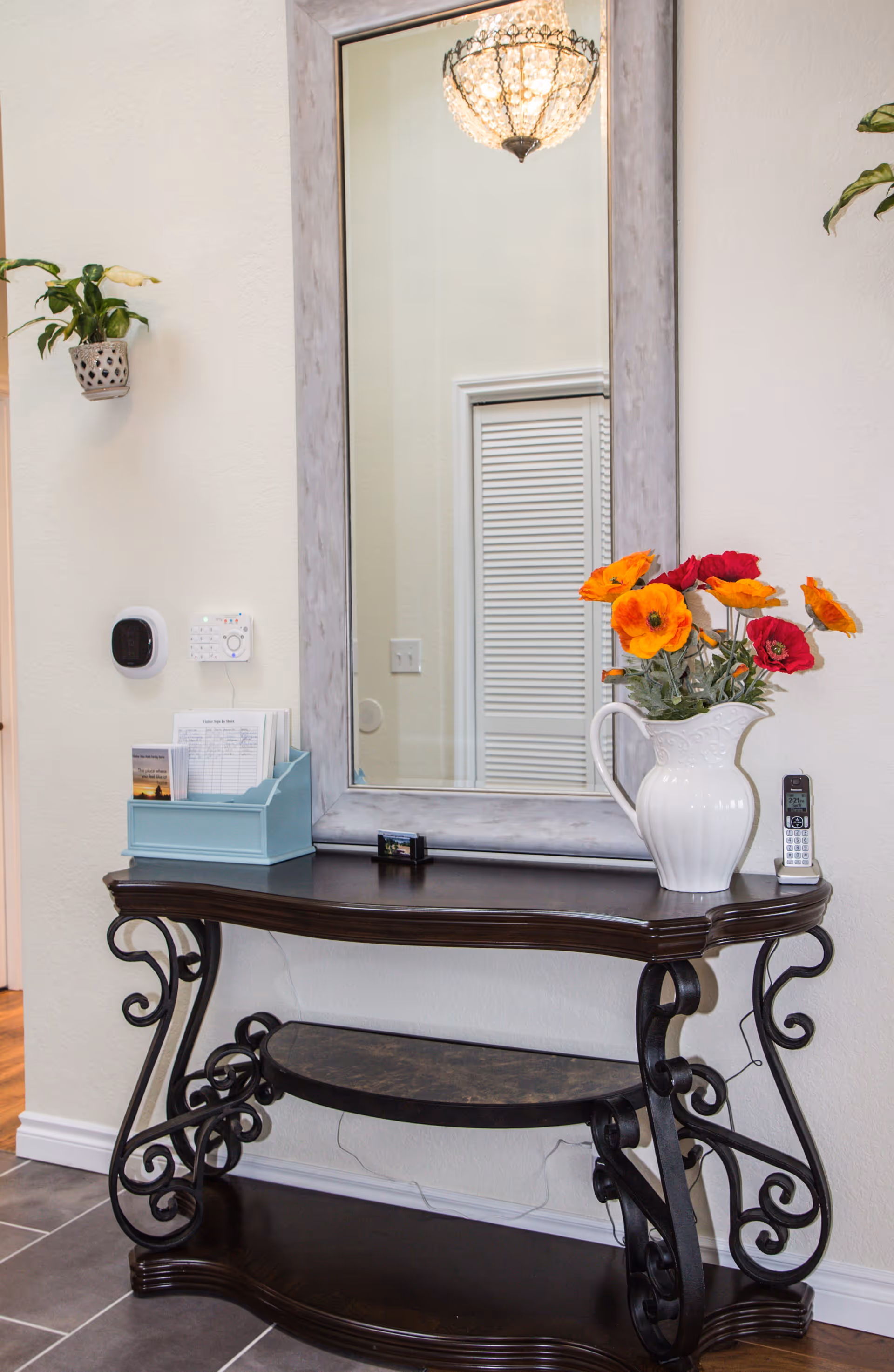 A decorative wooden console table with ornate black metal legs placed against a beige wall. On the table is a white pitcher vase with orange and red flowers, a cordless phone, a blue organizer holding papers, and a small framed photo. Above the table is a large rectangular mirror with a gray frame reflecting a ceiling light fixture and a white louvered door.