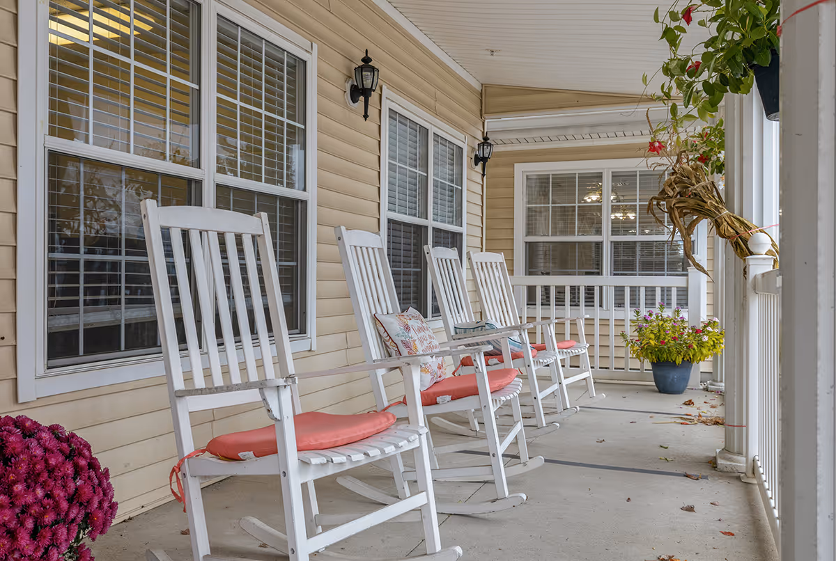 A covered porch area with four white wooden rocking chairs, each with a red cushion. The porch has beige siding, white-framed windows with blinds, two black wall-mounted lantern lights, and several potted plants including flowers and greenery. The porch floor is concrete with some scattered leaves.