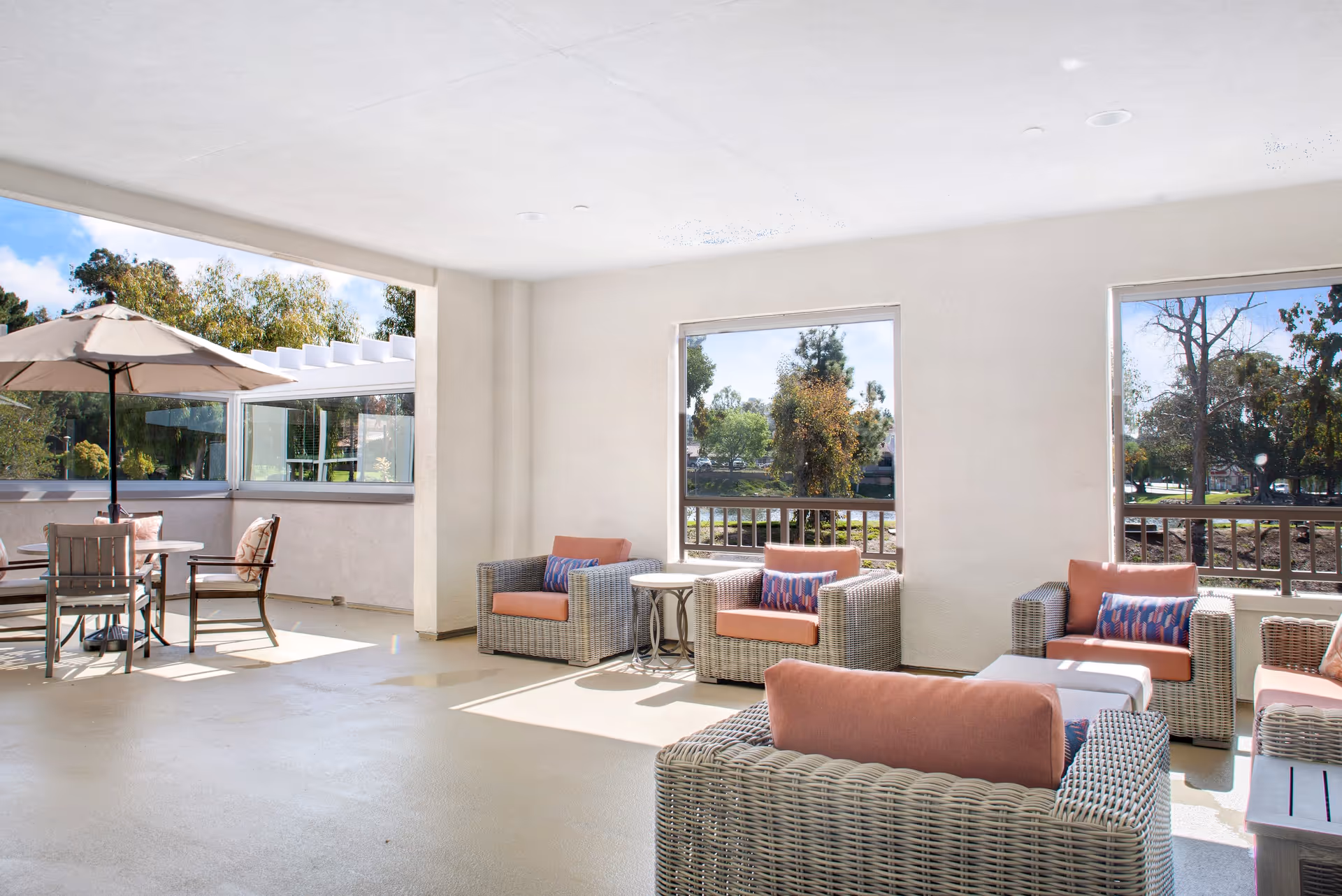 A bright and spacious outdoor patio area at Sunrise Of Orange featuring wicker armchairs with coral cushions and patterned pillows, a round table with chairs and an umbrella, and large windows offering views of trees and greenery outside.