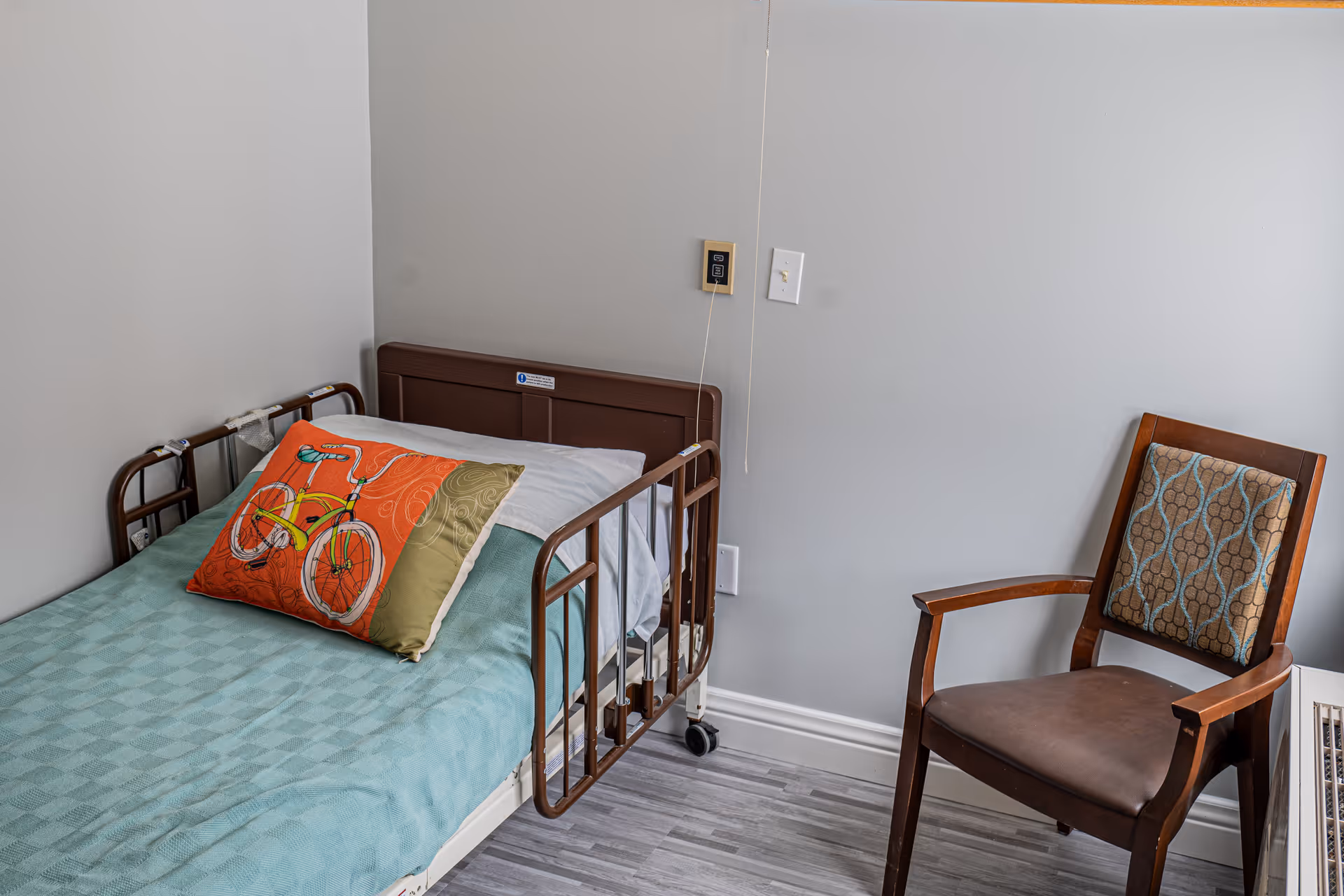A small room with a single hospital-style bed with brown rails, a teal blanket, and a decorative pillow featuring a yellow bicycle on an orange and green background. Next to the bed is a wooden chair with a patterned cushion. The walls are light gray and the floor has a gray wood-like pattern.