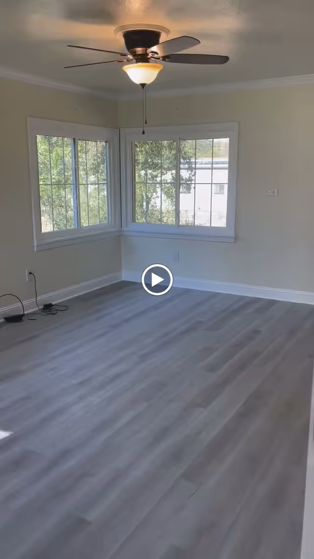 Empty room with light gray wooden flooring, beige walls, two large corner windows with white frames showing trees outside, and a ceiling fan with light fixture.
