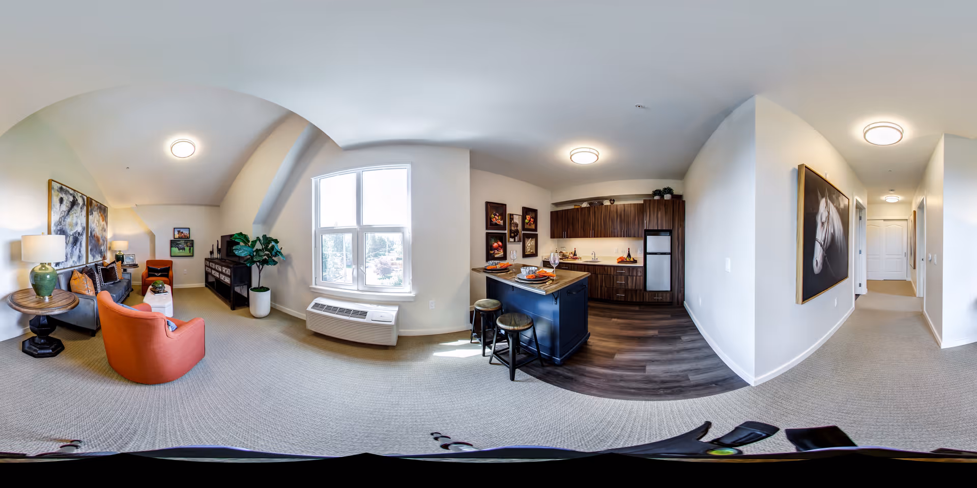 A bright and spacious senior living apartment interior featuring a living room with a sofa, orange armchair, side table with a lamp, and artwork on the walls. Adjacent to the living room is a kitchen area with dark wood cabinets, a small island with two stools, and a refrigerator. A large window lets in natural light, and a hallway with a horse painting on the wall leads to other rooms.