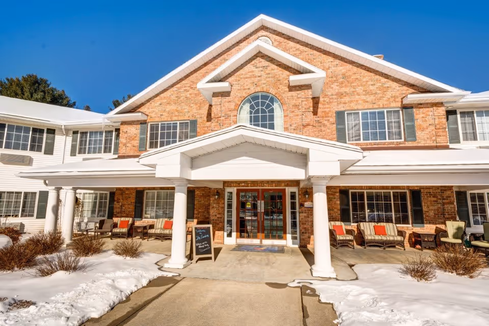 Front exterior view of The Landing at Queensbury facility with a brick and white siding facade, large windows, a covered entrance supported by white columns, outdoor seating with chairs and cushions, and snow on the ground under a clear blue sky.