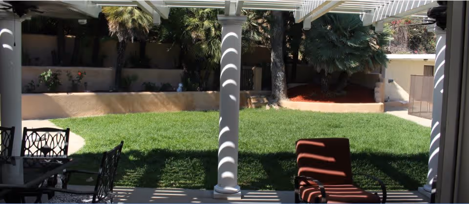 View of a shaded outdoor patio area with white columns and a pergola casting striped shadows. There is a red cushioned chair and a black metal table with chairs on a concrete floor, overlooking a green grassy yard with trees and plants along a beige wall.