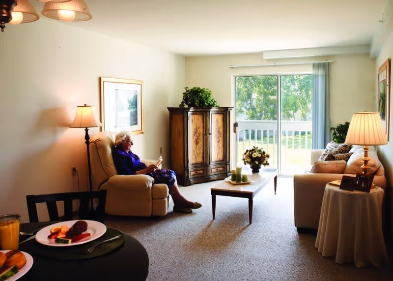 A cozy living room with an elderly woman sitting in a beige recliner chair reading a book. The room has a beige carpet, a wooden coffee table with candles and a flower arrangement, a beige sofa with pillows, a side table with a lamp and framed photos, and a wooden cabinet with a plant on top. Large sliding glass doors let in natural light and show a green outdoor area.