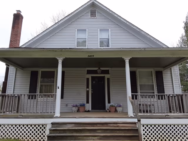 Front exterior view of a two-story house with a covered porch supported by two white columns, wooden railings, and steps leading up to a black front door. There are two windows on either side of the door and two windows on the second floor. Several potted plants are placed on the porch.