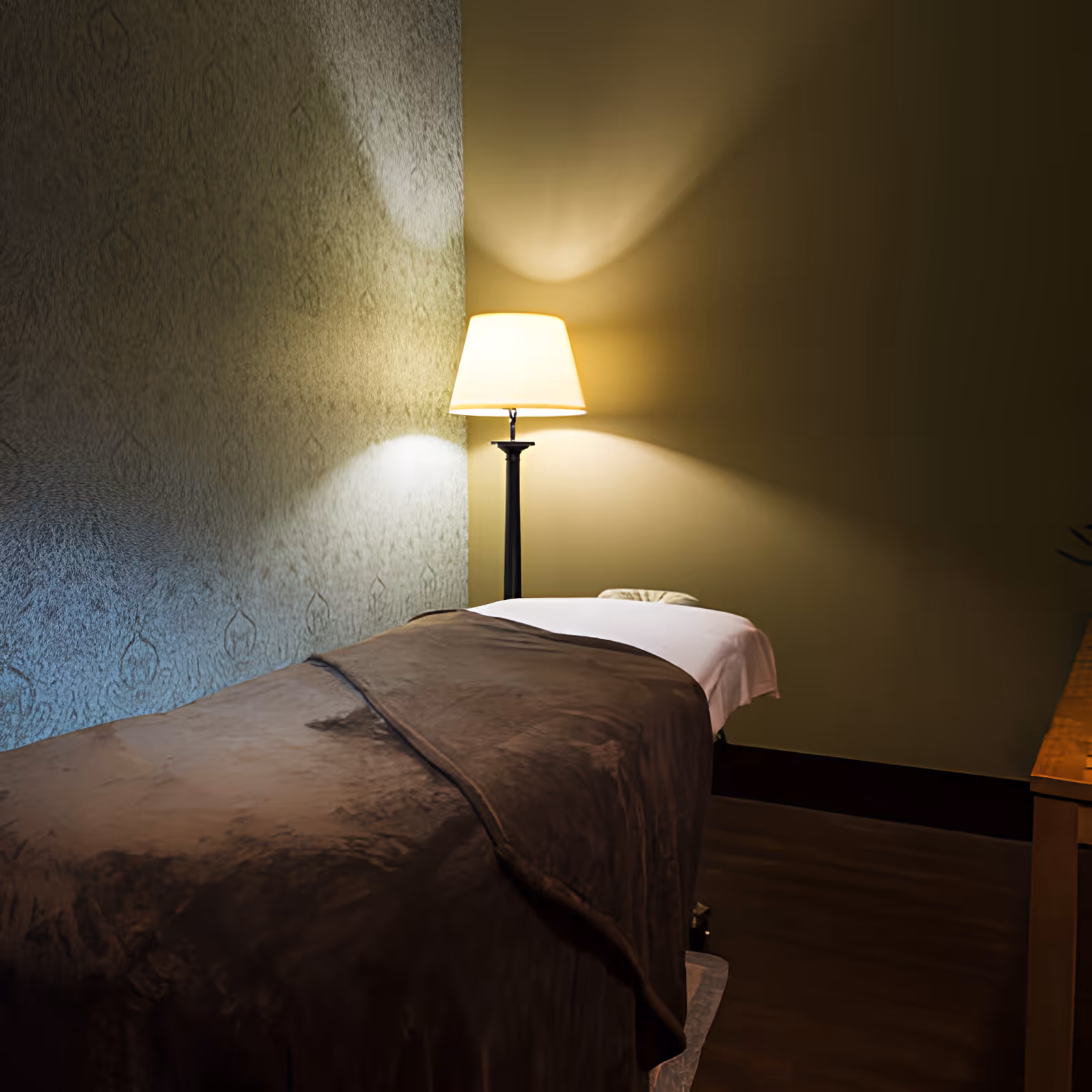 Dimly lit interior with a covered massage or treatment table and a floor lamp casting light on the wall.