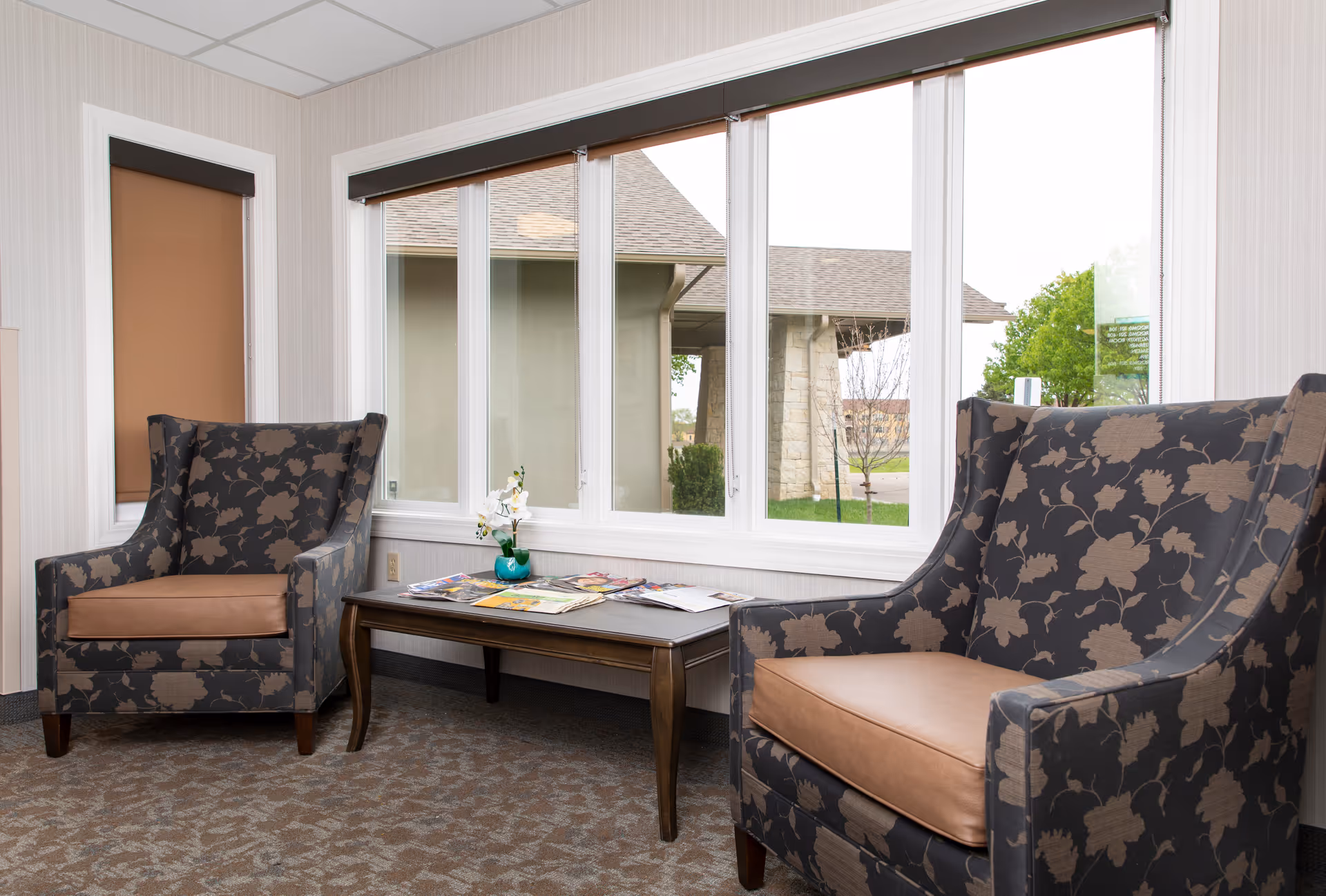 Two patterned armchairs flank a coffee table with magazines in a bright seating area by large windows.