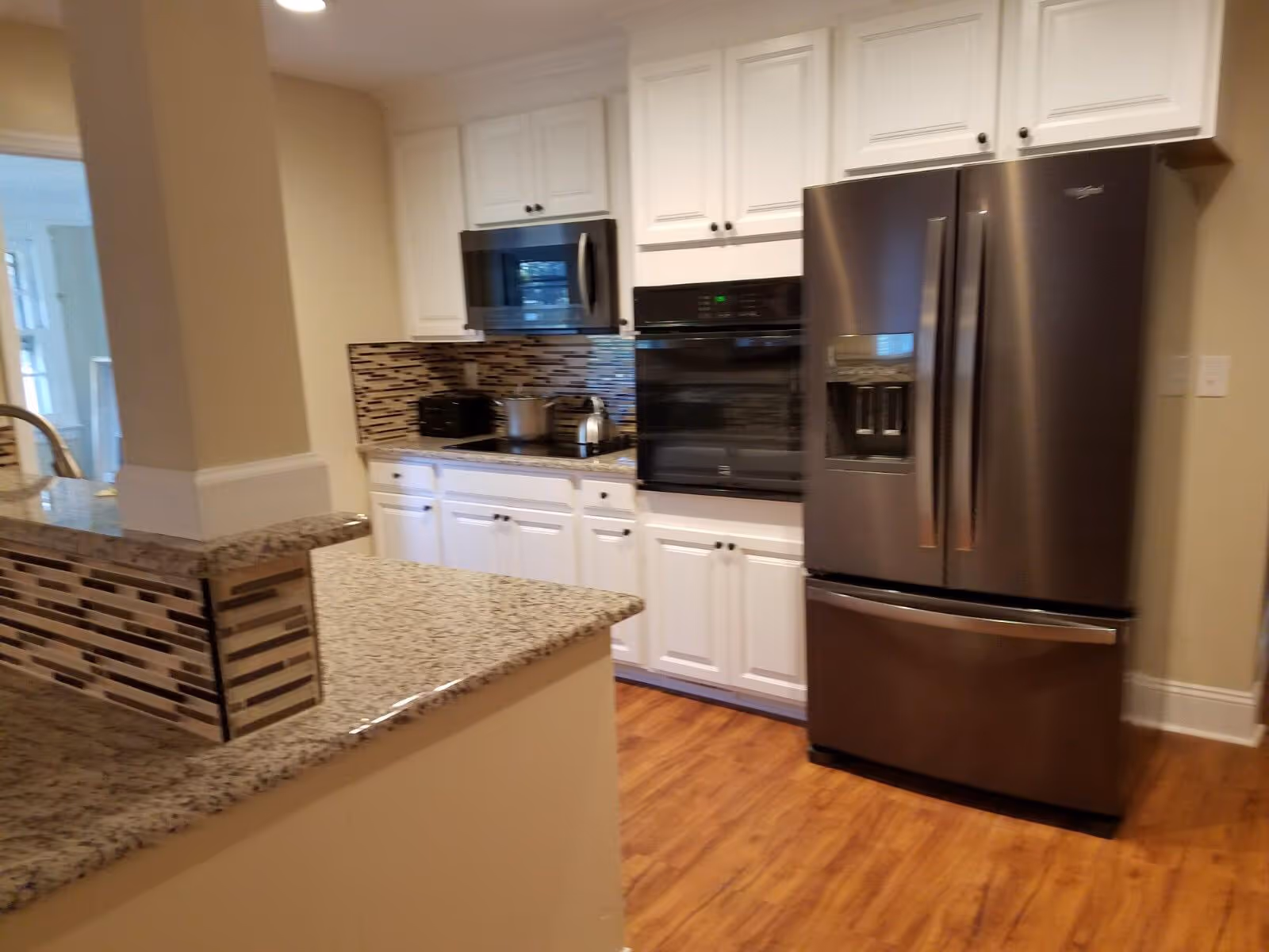 Modern kitchen with white cabinets, granite countertops, a stainless steel refrigerator with a water dispenser, a built-in oven, microwave, and a tiled backsplash. The floor is wooden and the kitchen has a clean, contemporary design.