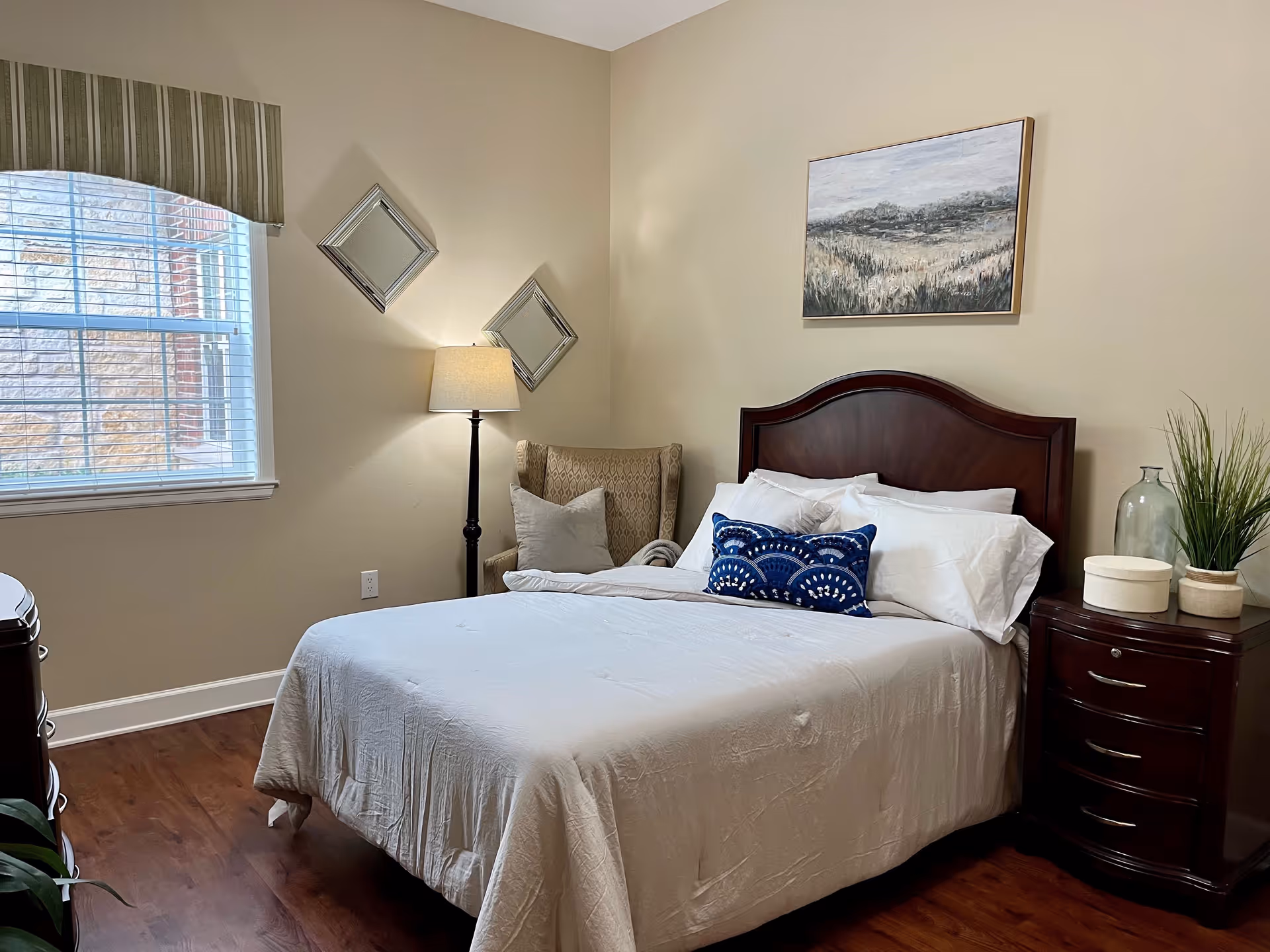 A cozy bedroom with a wooden bed frame, white bedding, and decorative blue pillows. There is a wooden nightstand with decorative items and a plant, a floor lamp, a cushioned armchair in the corner, two diamond-shaped mirrors on the wall, and a window with blinds and a striped valance. A landscape painting hangs above the bed.