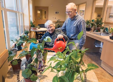 An elderly woman and an elderly man are watering various potted plants on a table inside a bright room with large windows. The woman is holding a red watering can and smiling, while the man stands beside her, also smiling. The room has a sink, cabinets, and additional plants on the counter.