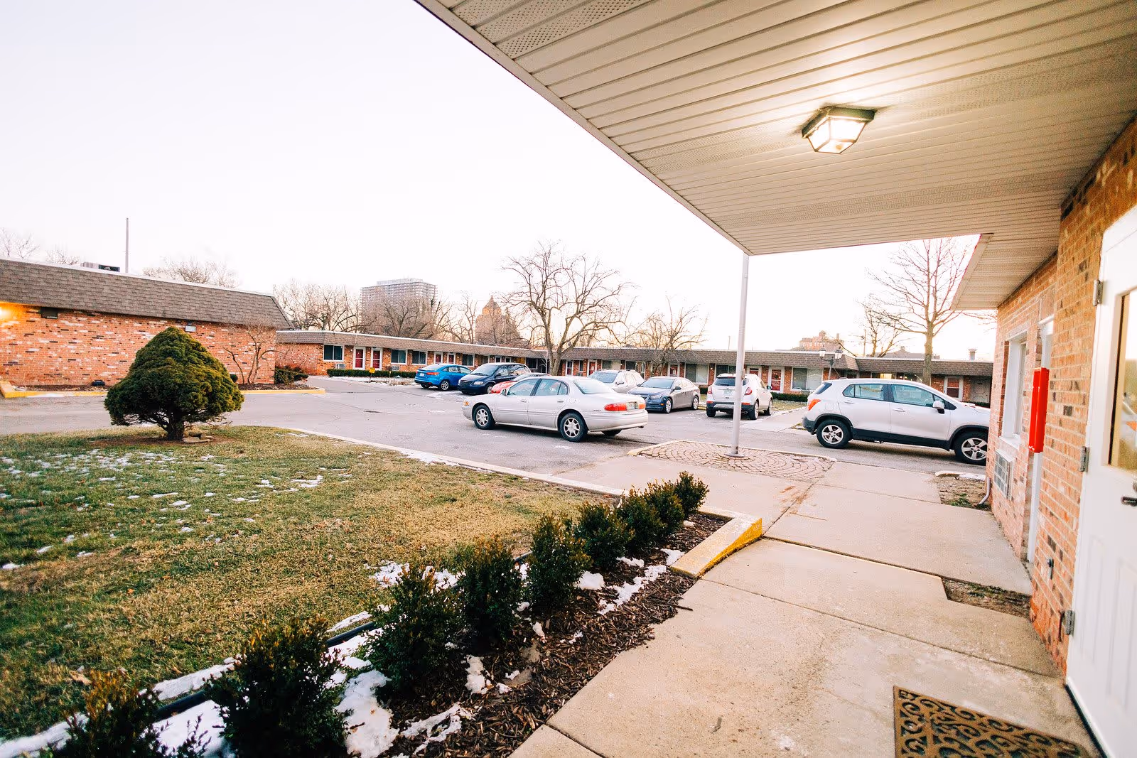 Outdoor view of a senior living facility parking lot with several parked cars, a small landscaped area with bushes and a tree, and a covered walkway attached to a brick building.