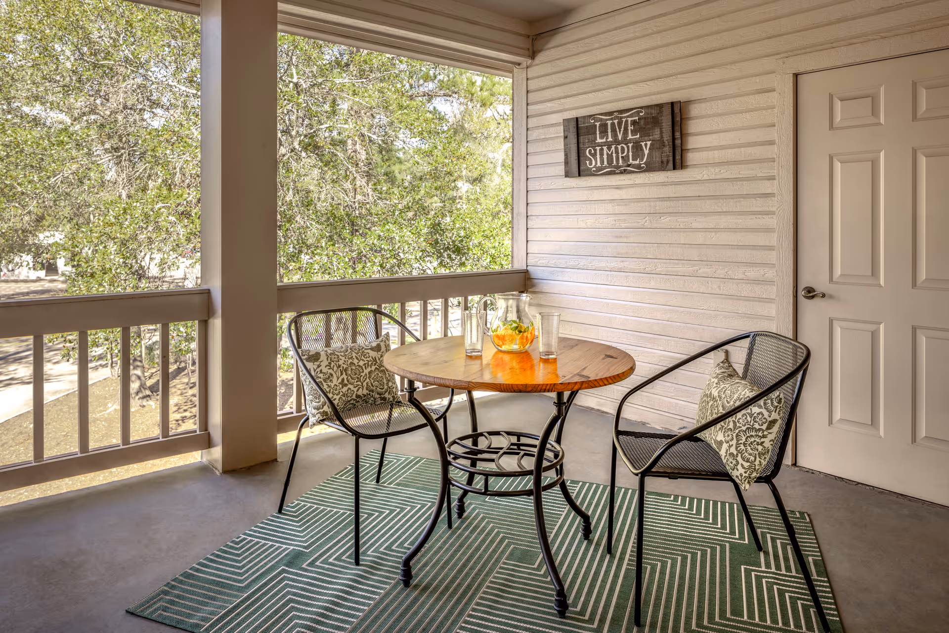 A covered outdoor patio area with a round wooden table and two black metal chairs with patterned cushions. On the table, there is a glass pitcher with orange slices and two empty glasses. The patio has a green geometric patterned rug, beige siding walls, a white door, and a decorative sign that reads 'LIVE SIMPLY'. Trees and greenery are visible outside the railing.