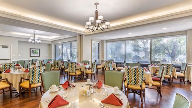 A bright and spacious dining room with multiple round tables set with white tablecloths, red folded napkins, glassware, and white cups. The room features large windows allowing natural light to fill the space, green cushioned chairs, wooden flooring, and elegant chandeliers hanging from the ceiling.
