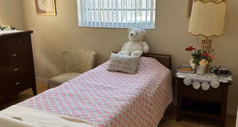 A small bedroom with a single bed covered in a pink patterned bedspread. A white teddy bear and a decorative pillow with text sit on the bed. There is a cushioned chair in the corner, a dark wooden dresser on the left, and a nightstand with a lamp, flowers, and books on the right. A window with vertical blinds is above the bed.