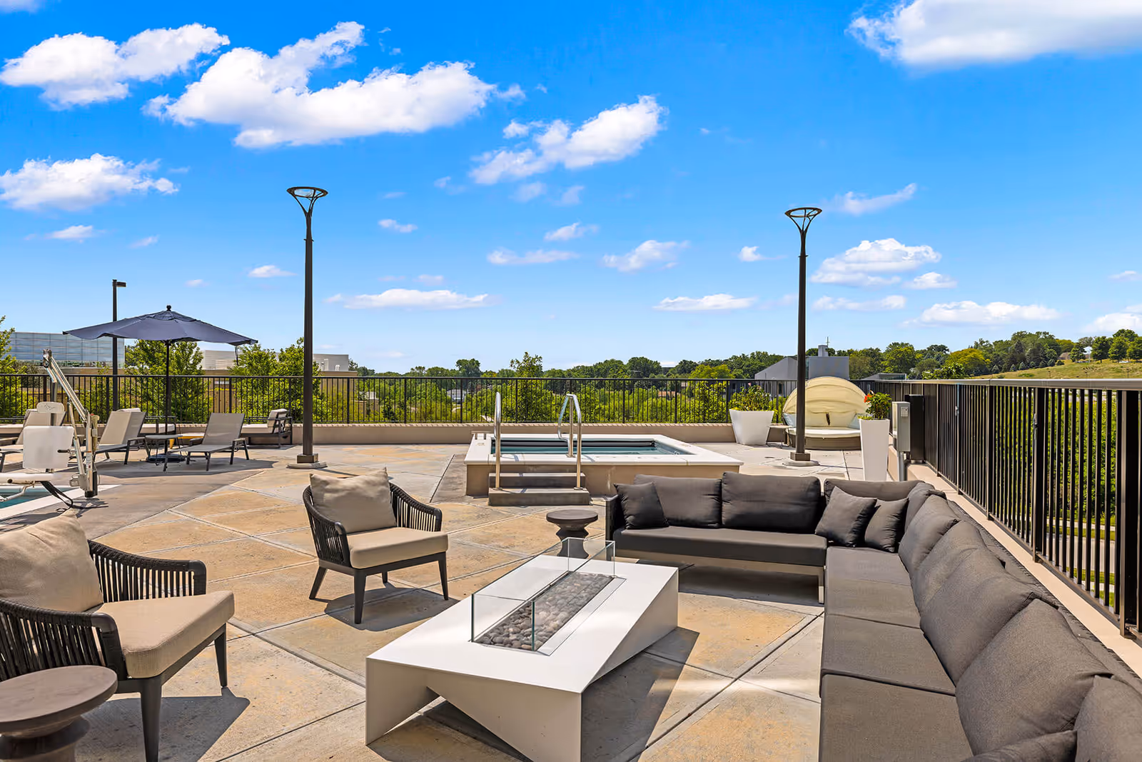 Rooftop outdoor lounge area with a sectional sofa, chairs, a fire pit table, loungers, umbrella, and a small pool/jacuzzi under a blue sky.