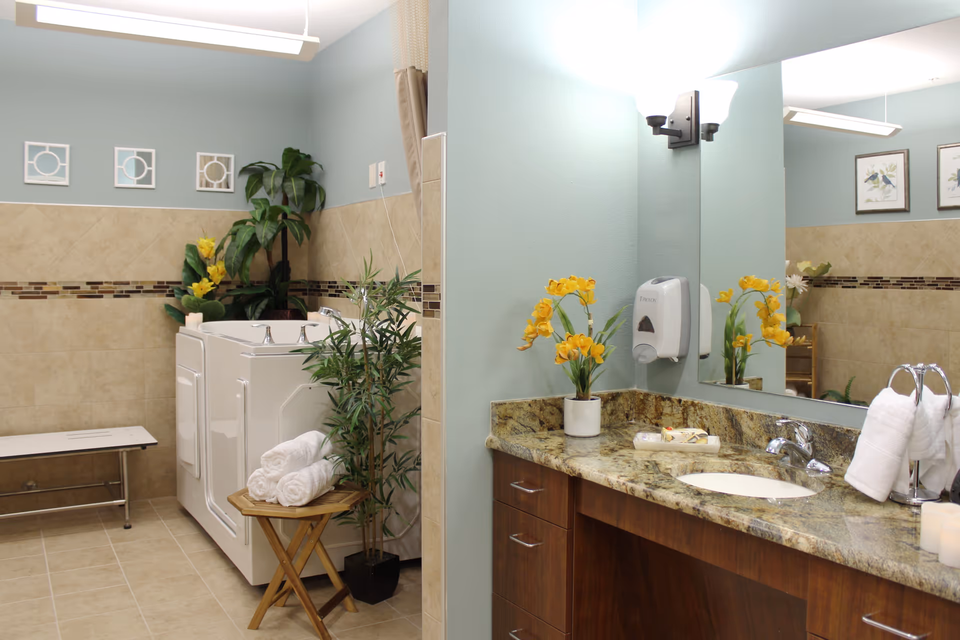A clean and well-lit bathroom featuring a walk-in bathtub with a side door, a bench, and a small wooden table holding rolled white towels. The walls are tiled halfway up with beige tiles and decorated with small framed art pieces and plants. A granite countertop with a sink, a soap dispenser, and yellow flowers in a vase is visible, along with a large mirror and wall-mounted light fixtures.