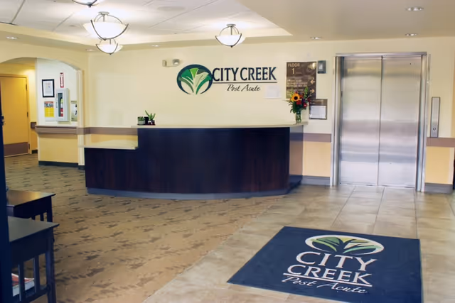 Reception area of City Creek Post Acute facility featuring a curved dark wood front desk, a wall sign with the facility's logo and name, an elevator with closed stainless steel doors, a floor mat with the facility's logo, and a hallway leading to other rooms.