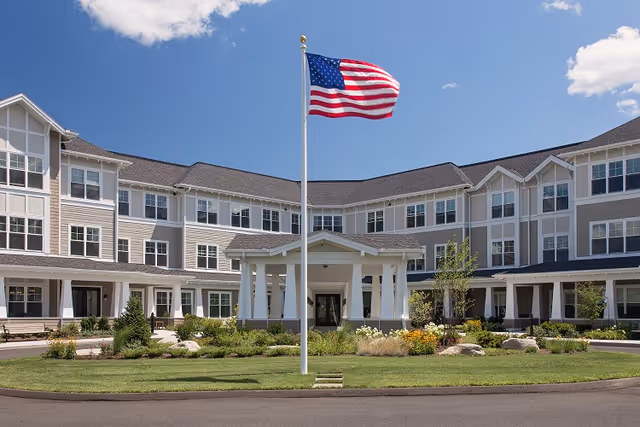 Front exterior of a multi-story senior living building with a flagpole flying an American flag in a landscaped circular driveway.
