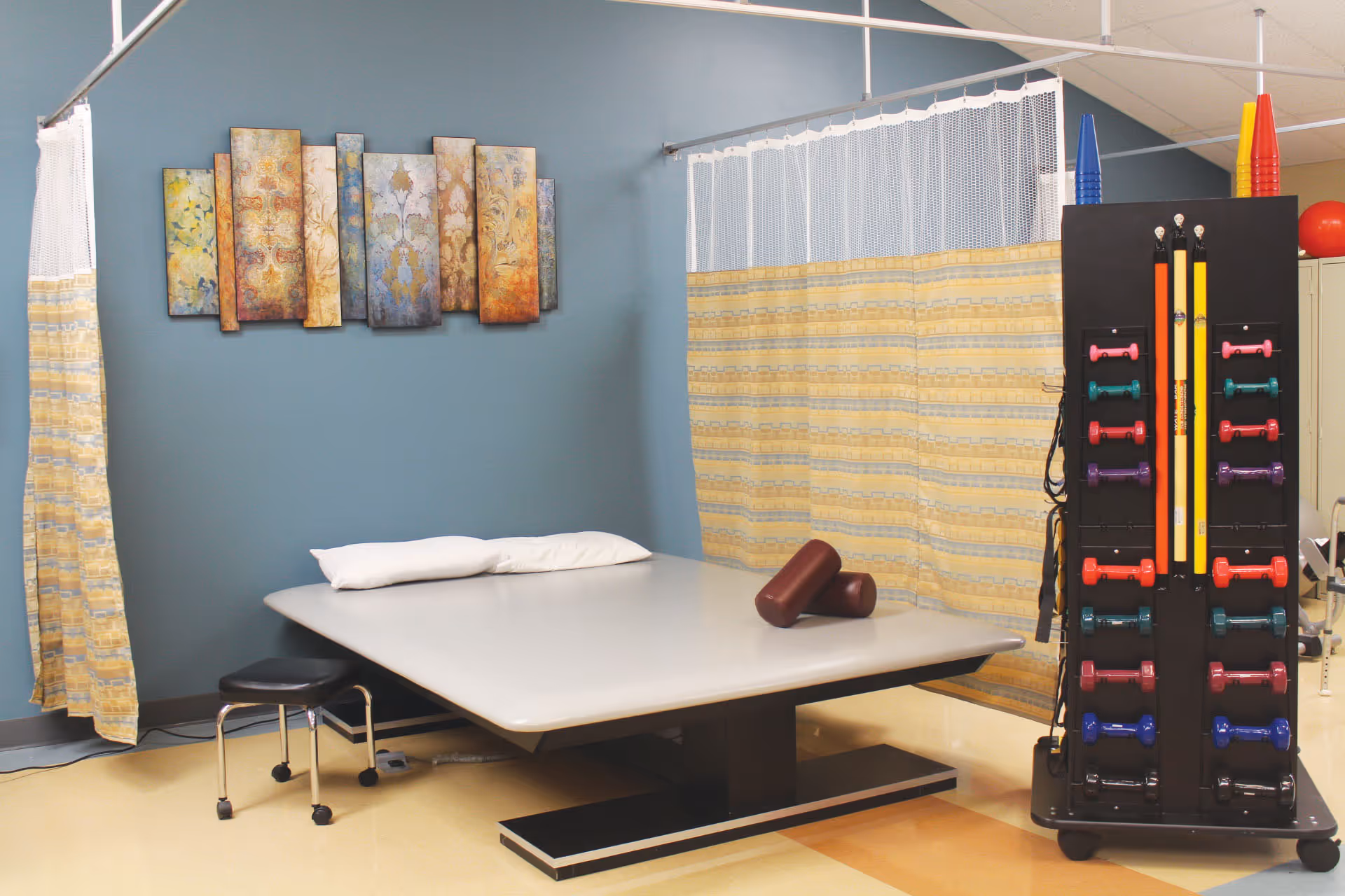 A therapy/rehab room with a treatment table topped by pillows, privacy curtains, wall art, and a rack of colorful hand weights and exercise equipment.