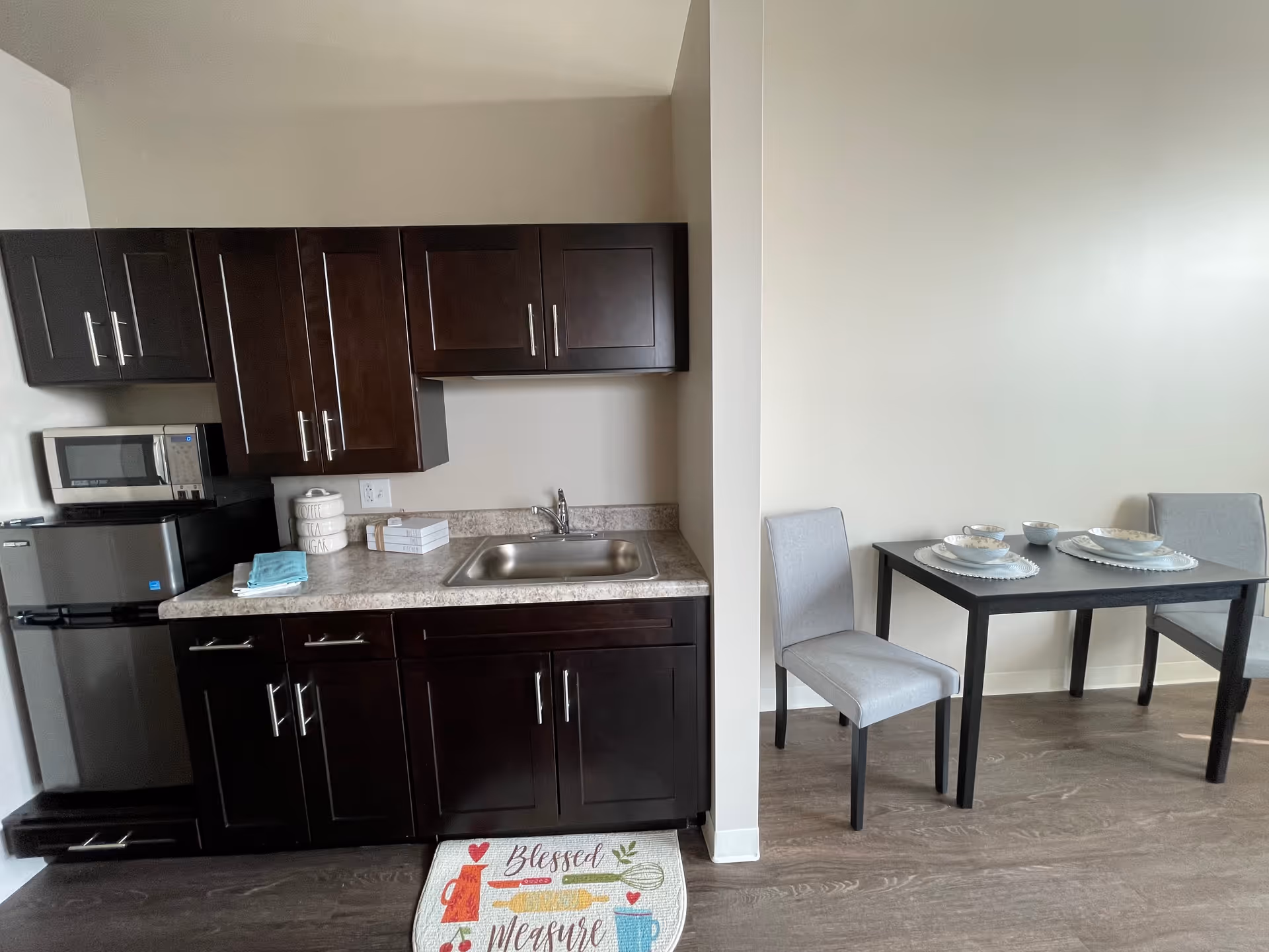 Small kitchen area with dark wood cabinets, a sink, mini fridge and microwave, and a two-seat dining table against a pale wall.