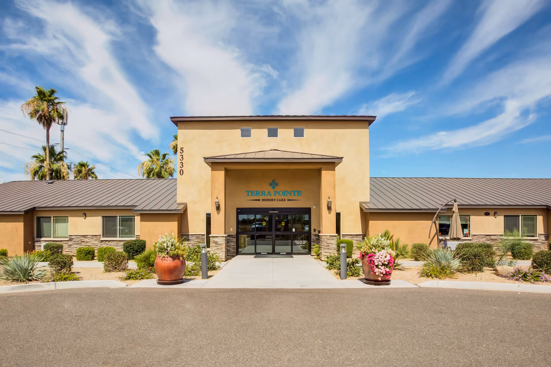 Front exterior view of Terra Pointe Memory Care facility with a clear blue sky and some clouds above. The building has a beige and stone facade with a metal roof, large entrance doors, and two large flower pots with plants on either side of the walkway leading to the entrance. Palm trees and shrubs are visible around the building.