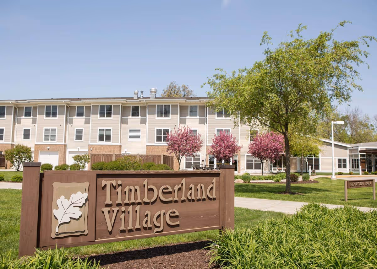 Exterior view of a senior living facility named Timberland Village with a large wooden sign in the foreground. The building is three stories tall with multiple windows, surrounded by green grass, trees, and flowering bushes under a clear blue sky.
