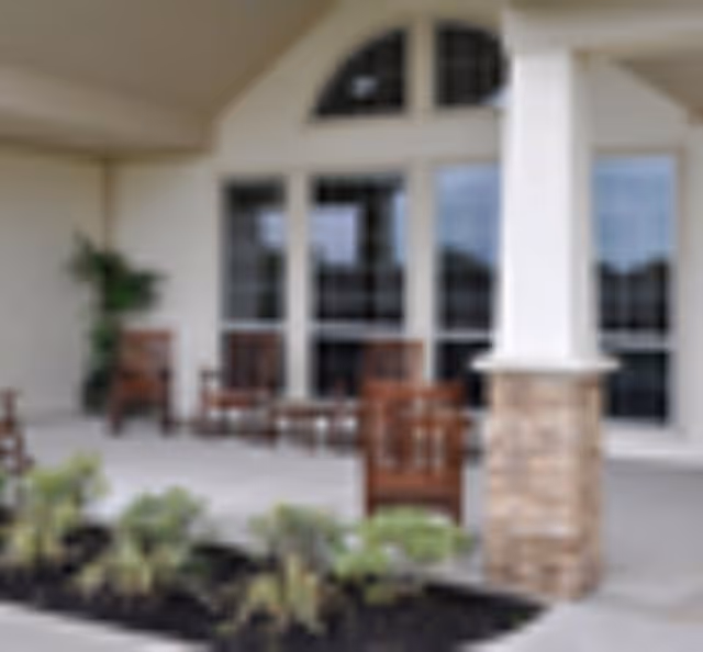 Covered front porch with wooden chairs, columns, large windows, and planted landscaping.