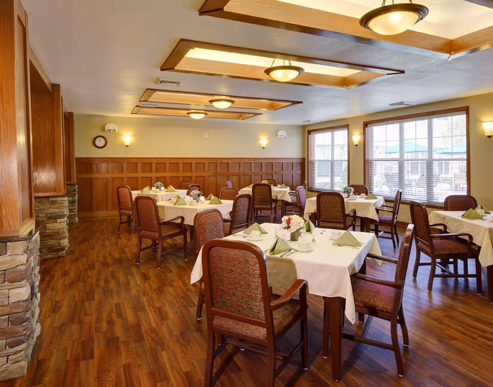 A senior living facility dining room with several tables covered in white tablecloths and set with green napkins, cups, and small floral centerpieces. The room features wood-paneled walls, large windows letting in natural light, and wooden flooring. Ceiling lights and wall sconces illuminate the space.