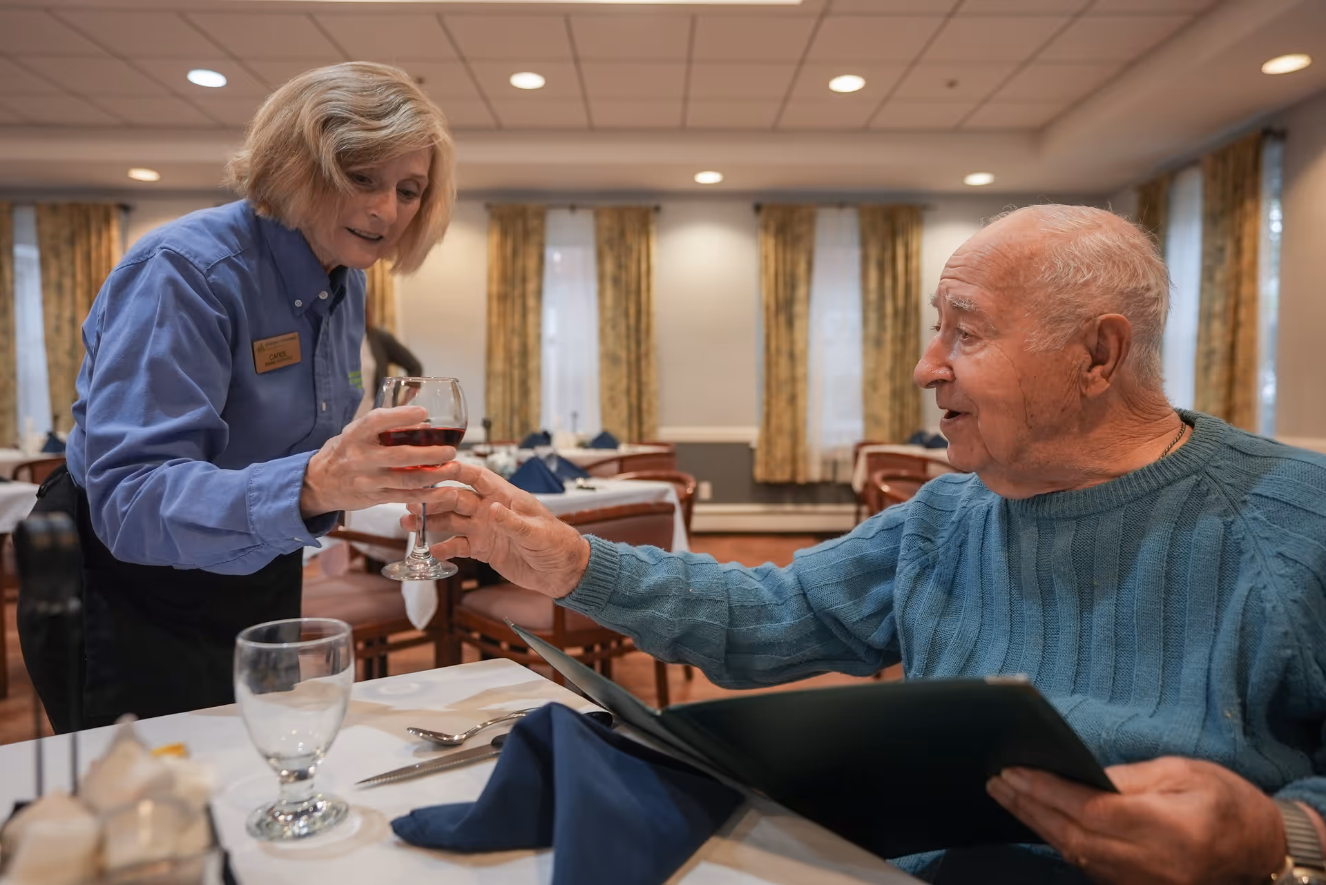 An elderly man in a blue sweater sitting at a dining table in a restaurant or dining room, reaching out to receive a glass of red wine from a female server wearing a blue shirt and a name tag. The room has multiple tables with white tablecloths, blue napkins, and wooden chairs, with windows and patterned curtains in the background.