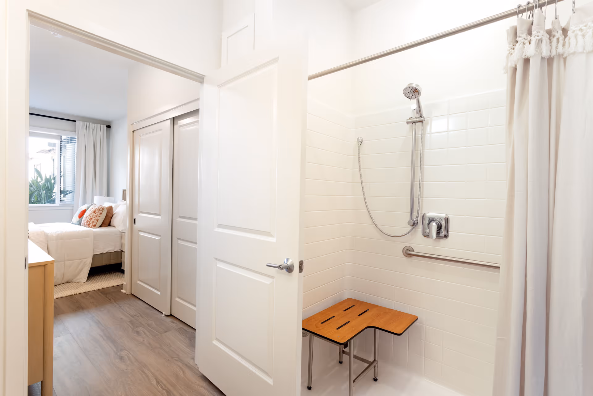 View of a bathroom with a walk-in shower featuring a wooden shower bench, grab bar, and a showerhead with a hose. The bathroom door is open, showing a glimpse of a bedroom with a bed, pillows, window, and curtains.