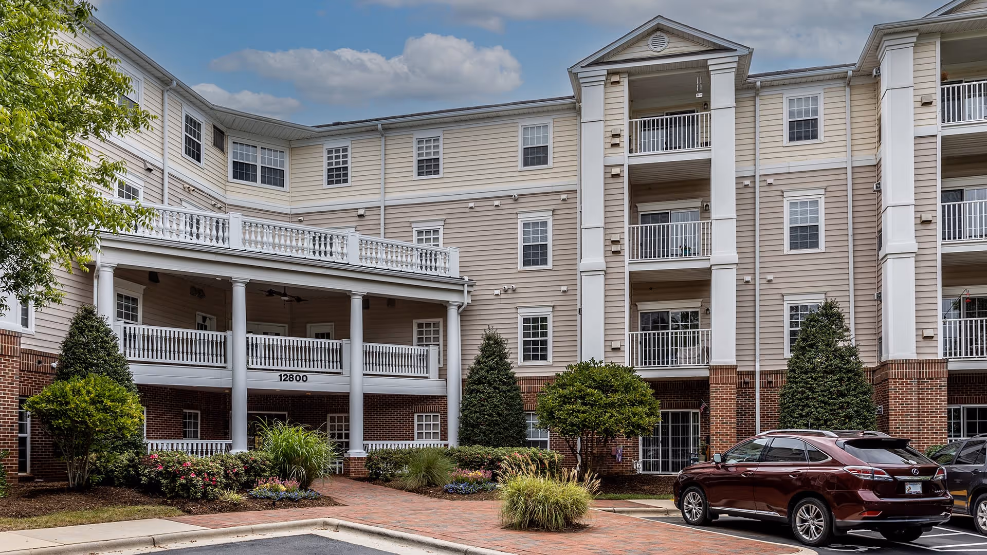 Exterior view of a multi-story senior living facility building with beige siding and red brick accents. The building features balconies, white railings, and a covered entrance supported by white columns. There are landscaped bushes and plants around the entrance, and a maroon SUV is parked in front. The building number 12800 is visible above the entrance.