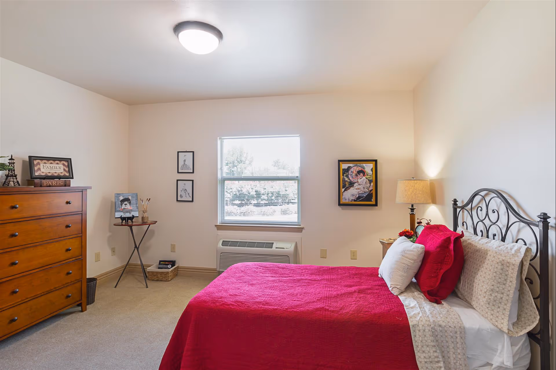 A cozy bedroom with a bed covered in a red quilt and white pillows, a wooden dresser on the left, a small round table with art supplies, and framed artwork on the walls. A window with blinds is centered on the back wall, and a lamp is on a nightstand next to the bed.