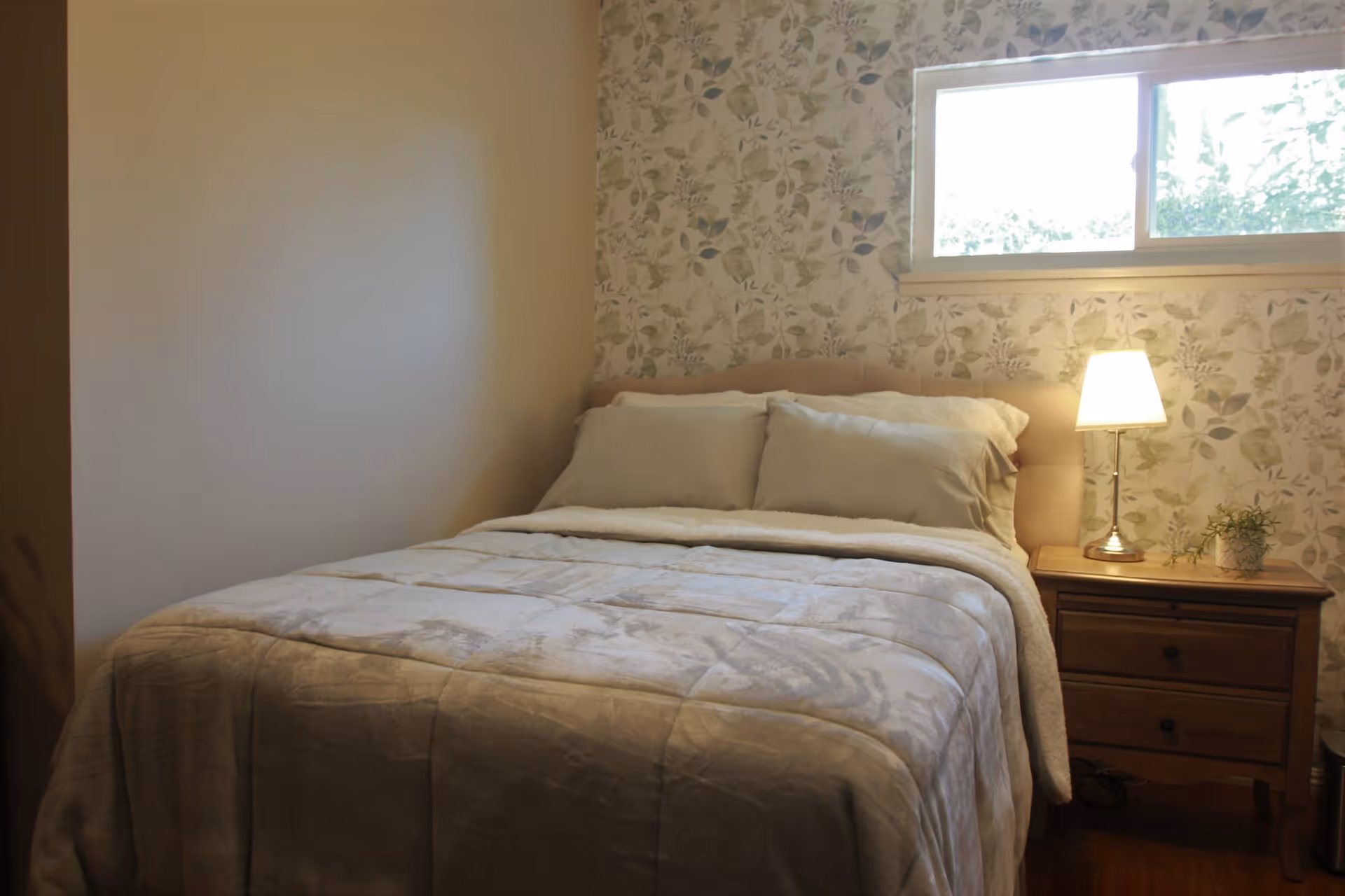 A neatly made bed with pillows and a lamp on a wooden nightstand beneath a window and floral wallpaper.