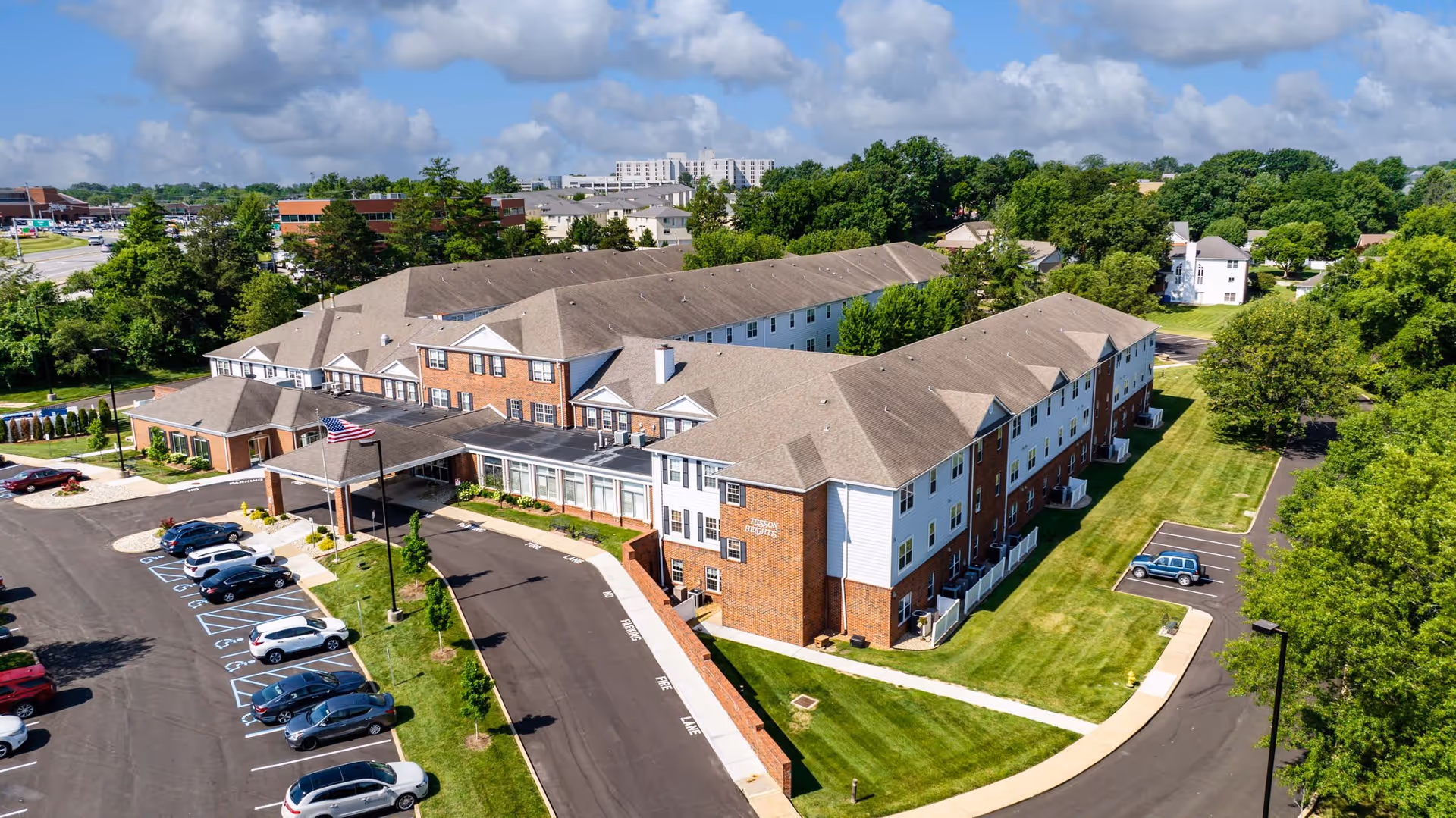 Aerial view of Cedarhurst Senior Living of Tesson Heights showing a large, two-story senior living facility with a brick and white exterior, surrounded by green lawns and trees. The building has a covered entrance with an American flag, and a parking lot with several cars parked. The sky is partly cloudy.
