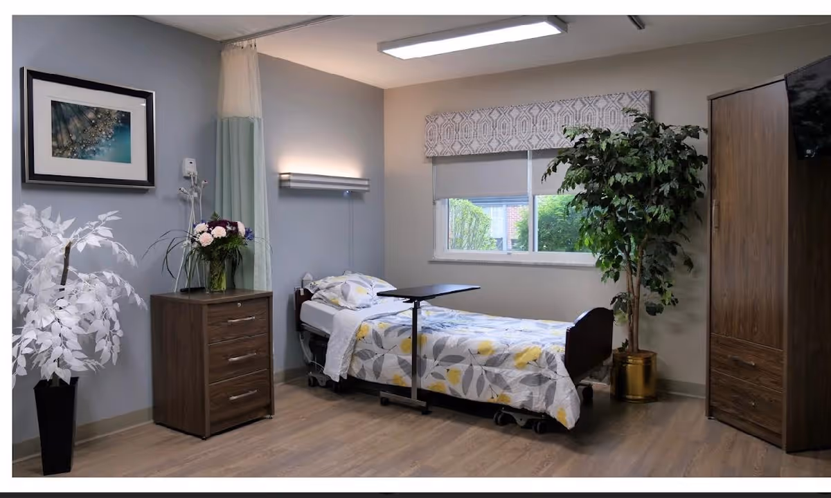 A neatly arranged nursing facility bedroom with a single bed covered in a floral patterned blanket. The room features a wooden dresser with a vase of flowers, a large potted plant, a window with a patterned valance, and a wooden wardrobe. The walls are painted in neutral tones and the floor has wood-like flooring.