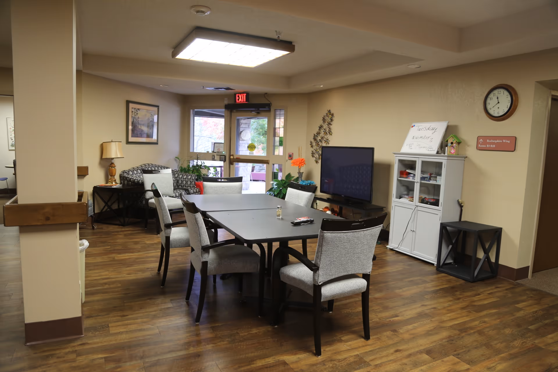 A common area in a senior living facility with a rectangular table surrounded by six cushioned chairs. There is a flat-screen TV on a stand against the wall, a white cabinet with glass doors, a clock, and a whiteboard with the date Tuesday, November 7. The room has wood flooring, beige walls, a couch with patterned cushions, a lamp on a side table, and an exit door with a window showing some greenery outside.
