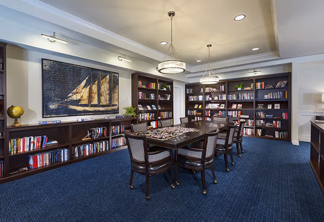 A cozy library room with dark wooden bookshelves filled with books along the walls. In the center, there is a large table with several chairs around it, and a puzzle is spread out on the table. The room has blue carpet flooring, two hanging ceiling lights, and a large painting of a sailboat on the wall.