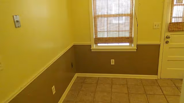 Empty interior corner of a room with tiled floor, two-tone painted walls in yellow and brown, a window with bamboo blinds, and a white door with a window also covered by bamboo blinds.