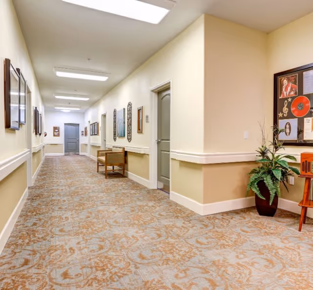 A well-lit hallway in a senior living facility with patterned carpet, beige walls, and several closed doors. The hallway is decorated with framed pictures and artwork on the walls, a wooden bench, and a potted plant near a bulletin board displaying various photos and documents.